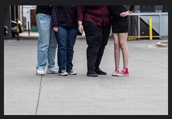 Four people, photo taken from waist down. Tall teen son loose light blue jeans, petite daughter in dark blue jeans, Mom with big Lipedema legs in black pants and teen daughter with short skirt and naked thin, knocked knee legs.
